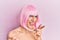 Young man wearing woman make up wearing pink wig pointing fingers to camera with happy and funny face