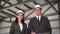 Young man and woman in helmets at a construction site. Businessmen in suits are looking at the camera and showing a