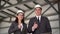 Young man and woman in helmets at a construction site. Businessmen in suits are looking at the camera and showing a
