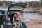 Young man and woman sitting in open trunk of car near the river and watching road map