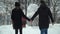 Young man and woman walking in winter park covered with snow holding hands. Winter leisure of happy loving couple. Back