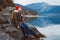 Young man with a yellow backpack wearing a red hat sitting on the shore on the background of mountain and lake. Space for your