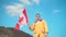 A young man in a yellow jacket, blue jeans and glasses stands in the mountains, holds the flag of Canada