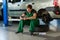 A young mechanic in a green overalls sits on a wheel in service