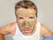 Young messy funny man looking at himself horrified in bathroom mirror with green cream on his face applying beauty facial mask pro