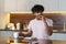 A young mestizo guy stands in the kitchen in subdued lighting, holds a mug in hand, eats a sandwich and drinks tea or