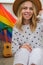 Young millennial hippie woman sitting on balcony using mobile phone. LGBTQ rainbow flag on background. Phone texting