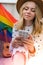 Young millennial hippie woman sitting on balcony using mobile phone. LGBTQ rainbow flag on background. Phone texting