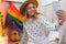 Young millennial hippie woman sitting on balcony using mobile phone. LGBTQ rainbow flag on background. Phone texting