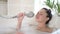 Young mixed race asian woman singing in bathtub using shower head. Happy millennial multi ethnic girl having fun enjoy