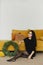 A young model woman in a black knitted dress sits near the yellow couch with a Christmas wreath in a minimalist interior