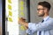 Young modern business man using adhesive notes while standing behind the glass wall in the office