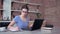 Young modern woman working at home, girl in eyeglasses typing on laptop keyboard and writes notes in clipboard sitting