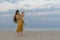 Young mom in bright yellow sundress and long hair holds baby. Portrait of mother with baby on sea background