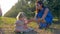 Young mom gives glass of water to cute toddler sitting on grass between rows of trees outdoors during apple crop season