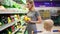Young mother is choosing yellow tomatos and other vegetables, while her little child is sitting in a grocery cart