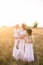 A young mother with her daughters with blond hair in white dresses at sunset in the summer in a field of countryside.