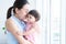 Young mother holding and kissing her cute little Caucasian 7 months newborn baby on her hands, standing near window at