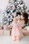 Young mother in pink dress with her little one year old daugter in cute dress preparing to the Christmas tree at home.