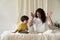 Young mother reading fairytales to little kid sitting on bed in bedroom entertain child on holiday