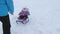 Young mother rolls little baby on sled along snowy road in winter. Child is naughty and cries while sitting in sled