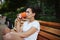 Young multi-ethnic woman drinking takeaway coffee from a disposable paper mug, sitting on wooden bench in a summer park