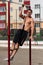 Young muscular athlete doing pull-up exercises hanging with one straight hand on a horizontal bar in the courtyard.