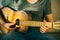 A young musician in a green t-shirt and blue jeans plays an old acoustic guitar