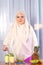 A young Muslim woman in a light scarf is standing at the table with fruit and putting kiwi puree into a glass.