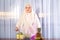 A young Muslim woman in a light scarf is standing at the table with fruit and putting kiwi puree into a glass.