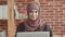 A young muslim woman, sitting in front of a laptop screen, looks up and looks at the camera . a girl in a hijab is