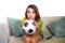 Young nervous and beautiful football fan woman watching television game sitting on sofa couch holding soccer ball excited highly c