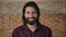 Young nice brunette man with beard is watching at camera, giggling, brick background