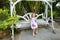 Young nice little girl riding on swing in exotic garden, palms in background.