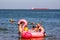 Young and old men ride small children on an inflatable pink flamingo and other vacationers and tourists swim in the sea