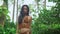 Young oriental model in brown crocheted bikini goes on path amid tropical trees