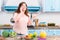 young overweight woman with wooden spoon in hand talking on smartphone while standing at table with fresh vegetables in kitchen