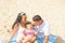 Young Parents Cute Laughing Baby Toddler Daughter with Kiss Mark on Cheek Sitting on Beach Smiling Looking at Each Other. Summer