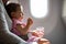 A young passenger infant sitting in an airplane wearing a seat belt in his mothers arms is preparing for his first trip