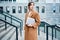 Young pensive casual businesswoman in coat with laptop dreamily looking away outdoor