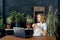 Young pensive dreaming woman with a cup of coffee and a laptop at the tables in the interior with plants in pots.