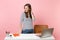 Young pensive woman looking down work and standing near white desk with contemporary pc laptop isolated on pastel pink