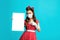 Young pinup woman in anti covid face mask pointing at blank paper poster with mockup for your design on blue background