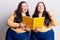 Young plus size twins holding book pointing thumb up to the side smiling happy with open mouth