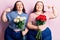 Young plus size twins holding flowers pointing finger to one self smiling happy and proud