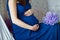 Young pregnant woman in a blue dress holds her hands on her swollen belly, love concept, violet flower