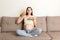 Young pregnant woman enjoys eating croissant on the sofa. Unhealthy pastry during pregnancy concept