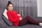Young pregnant woman lies on sofa in living room, looks at camera, touches her stomach through red warm sweater, looks peaceful,