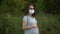 A young pregnant woman in a medical mask stands in nature. The girl in glasses and a dress looks into the camera.