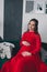 Young pregnant woman in red dress sitting in sofa in studio baby clothing